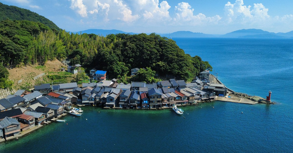 伊根灣沿岸的舟屋群空拍，一整排木造房屋的一樓緊貼水面，門前繫著漁船，岸端有一座紅色小燈塔