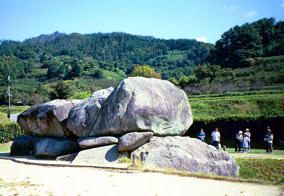 石舞台古墳:兩塊巨大的灰色花崗岩壓在下層石塊上、中間圍出一個石室的開口,前方是沙土步道,右側站著一小群遊客當比例尺,背後是階梯狀的綠色農地與整片森林覆蓋的山丘