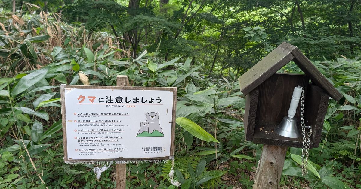 秋田市郊外林緣的日英雙語「クマに注意しましょう」告示牌，列出不要單獨行動、帶鈴出聲、不要靠近小熊、慢慢後退等五條建議，右側立著一座木造小屋形狀的鐘架，裡面掛著一只手搖鈴