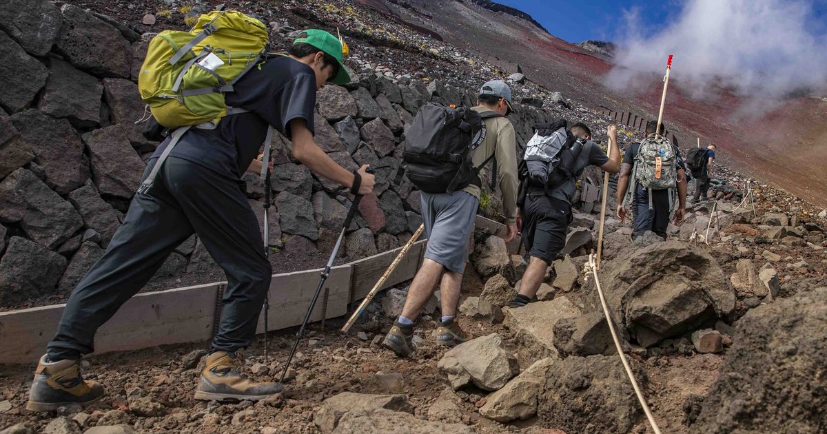 一列背著大背包、手持登山杖與木製金剛杖的登山者，沿著砌石護坡旁的碎石陡坡往上爬，右側拉著一條供人抓握的粗麻繩