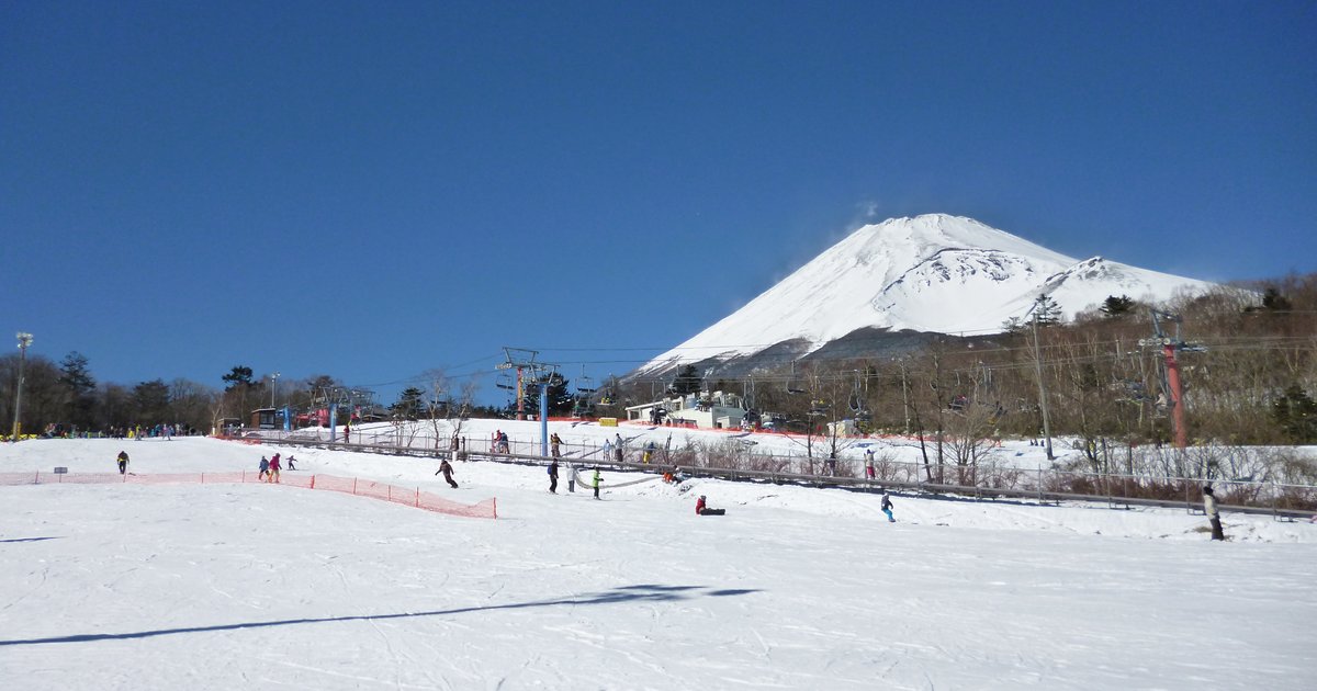 富士山 Yeti 滑雪場山腳平緩的雪坡,晴天藍空,右後方是整座積雪的富士山,中間幾座吊椅支柱與纜線往山上延伸,雪坡上有零星滑雪的人;2015 年 2 月雪季中段拍攝,當時名稱是スノータウン Yeti