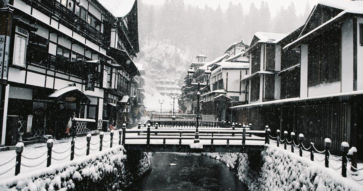 下雪中的銀山溫泉街：銀山川兩岸是三、四層樓的木造旅館，河上橫著一座木橋，岸邊的鐵鍊欄杆柱與煤氣燈都頂著一層雪，路面全是雪、看不到任何車輛