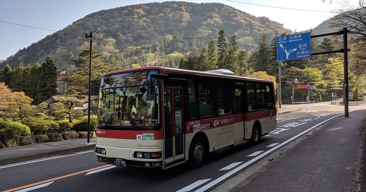 一輛紅白配色的箱根登山巴士行駛在箱根山區的雙線道上,車身側面印著 Hakone Tozan Bus,右方藍色路標上排寫著沼津 Numazu 與元箱根 Motohakone 並配国道 1 號的盾形標誌,下排是湖尻 Kojiri 與大涌谷 Owakudani,背景是整片森林覆蓋的山坡