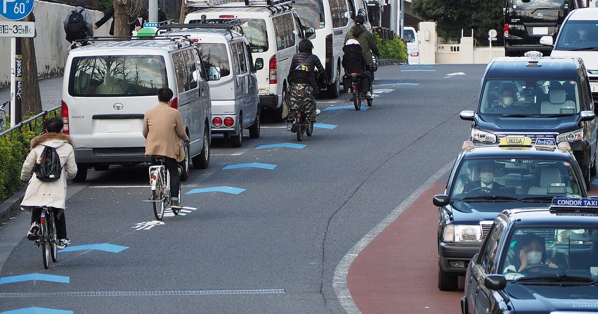 東京原宿的市區道路,四名騎士沿著車道左緣的藍色人字形自転車ナビライン 往前騎,左側停著一排白色廂型車,右側對向車道有計程車迎面駛來