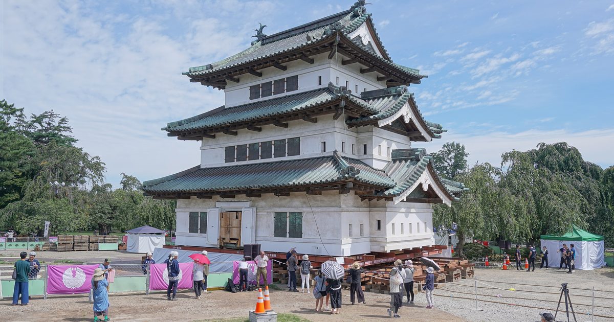 弘前城天守被整棟頂起、放在紅色鋼梁與木製枕木上,站在礫石地面上,四周圍著印有津輕家家紋的紫色布幕與圍觀民眾