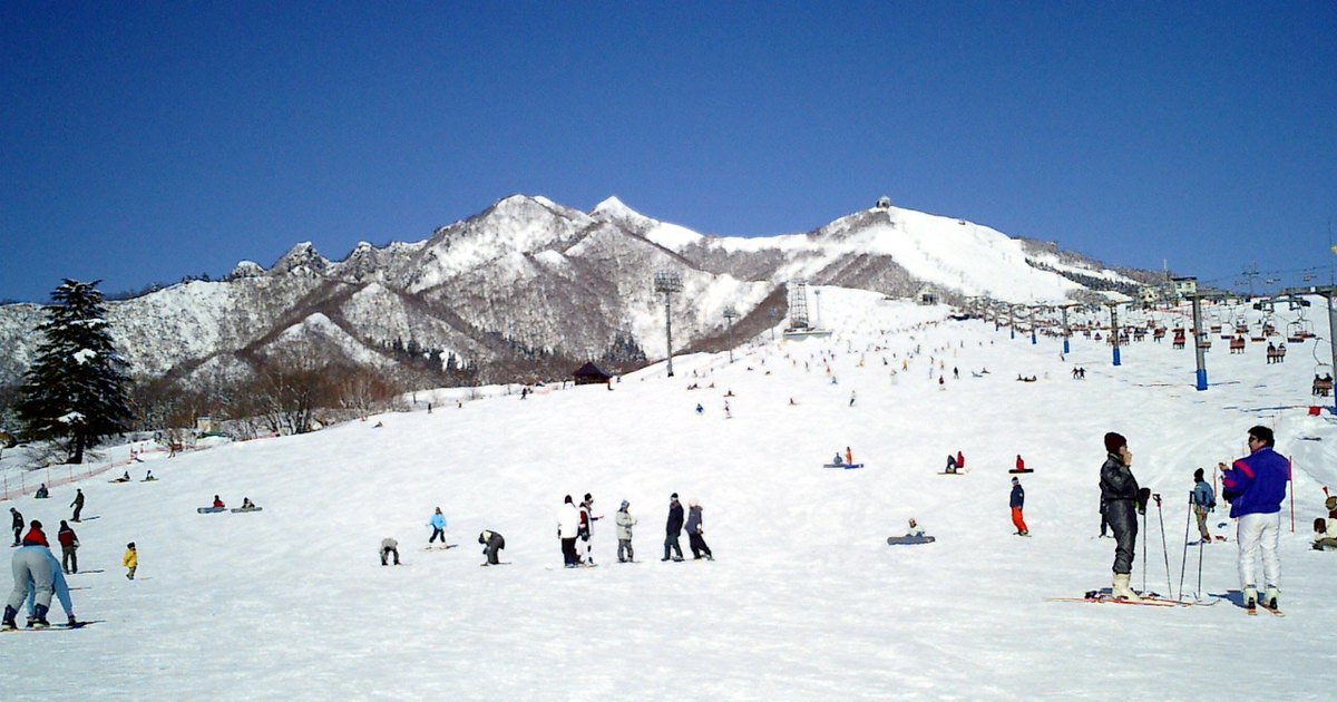 晴天的岩原滑雪場山腳：寬闊平緩的壓雪雪道往上延伸，右側一整排吊椅纜車，坡上立著高高的照明燈柱，後方是覆雪的飯士山一帶山稜；雪道上散著滑雪和單板的人。2004 年 2 月拍攝