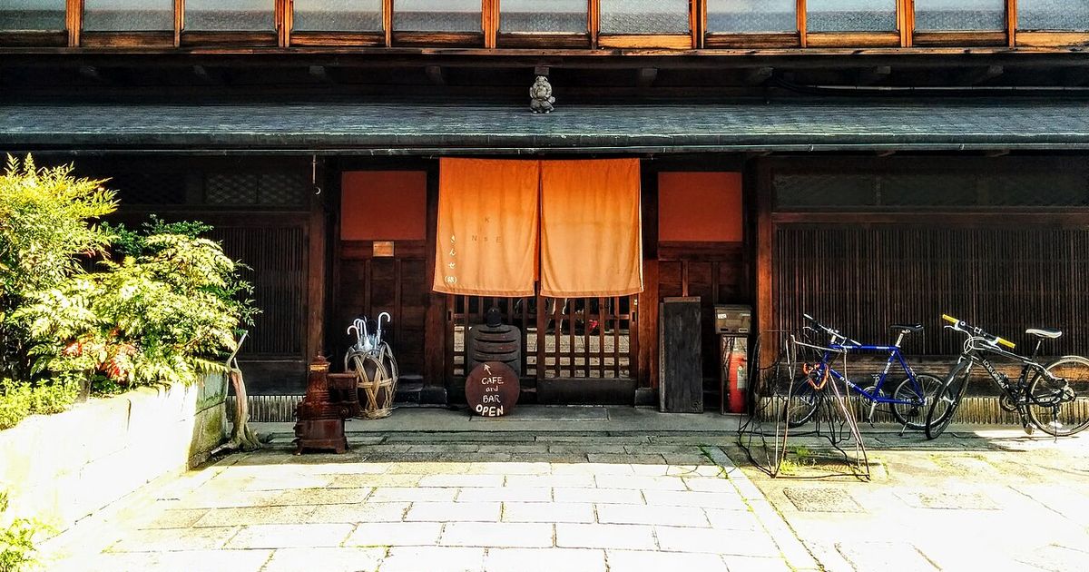 京都町家旅館的木造外觀:二樓一整排格子玻璃窗,一樓入口垂著橘色暖簾,門旁立著寫有 CAFE and BAR 的圓形黑板,右側停著兩台腳踏車,前方是石板路