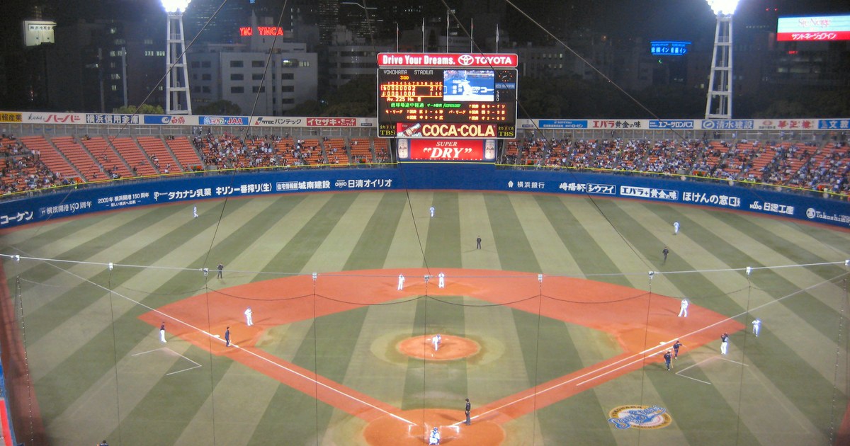 日本職棒球場的夜間比賽全景:內野紅土與條紋外野草皮在強力照明下相當明亮,看台上坐著觀眾,遠處記分板亮著