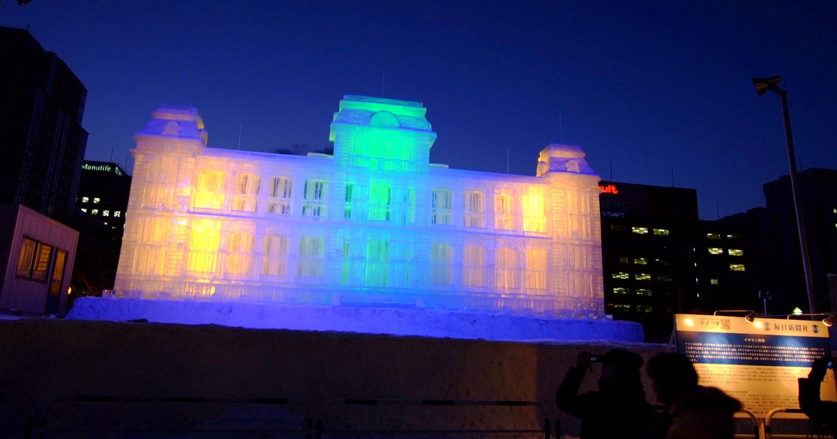 札幌雪祭大通會場夜間的大型冰雕,仿西式宮殿造型,打上黃、綠、藍色燈光,背景是深藍夜空與辦公大樓