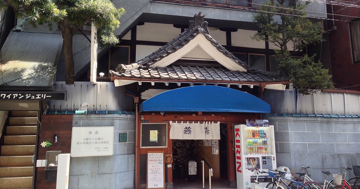 東京上野的錢湯「燕湯」外觀，瓦片唐破風屋簷下掛著白色暖簾、藍色遮陽棚，門邊立著寫有營業時間的看板
