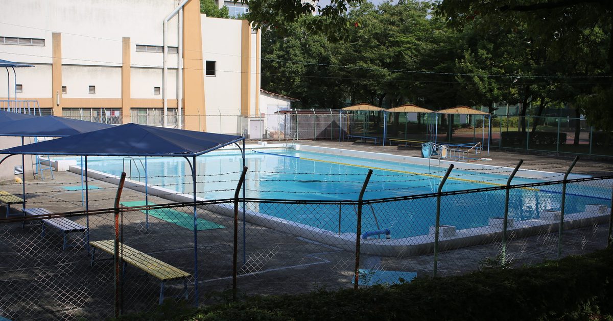 日本市營戶外泳池的夏季景象:長方形水池裝滿水、水面平靜無人,池畔架著三頂藍色遮陽棚與幾張長椅,右後方立著四把茅草頂遮陽傘,整區外圍是鐵絲網圍籬,背景為米黃色的體育館建築與樹林