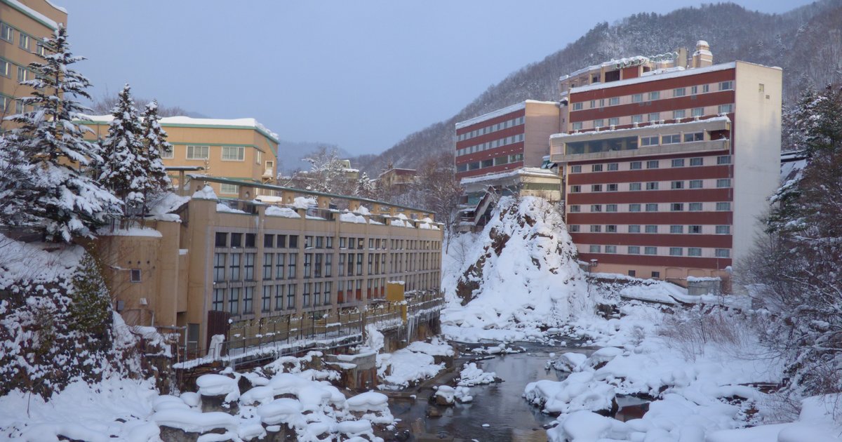 冬天清晨的定山溪溫泉:豊平川的溪石上堆滿積雪,兩岸是米黃色與紅白條紋的飯店建築,背後是覆雪的山坡