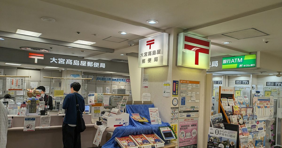 日本大宮髙島屋郵便局的營業廳,兩面紅色〒標誌的燈箱招牌下是長條櫃檯,一名穿深色制服的行員正在接待一位背側背包的顧客,中央桌面攤著印刷品與一個 ゆうパック 紙箱,右側是ゆうちょ銀行的 ATM 區與整排文宣架