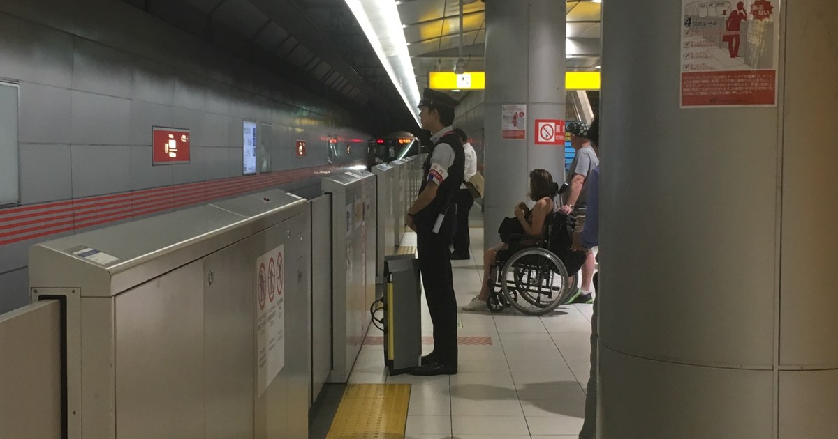 地下車站月台:戴制服帽的站務員站在月台門旁、手邊備著攜帶式斜坡板,另一側一名坐手動輪椅的乘客與同行者在柱子邊等車;地面是黃色導盲磚,隧道深處可見列車頭燈接近