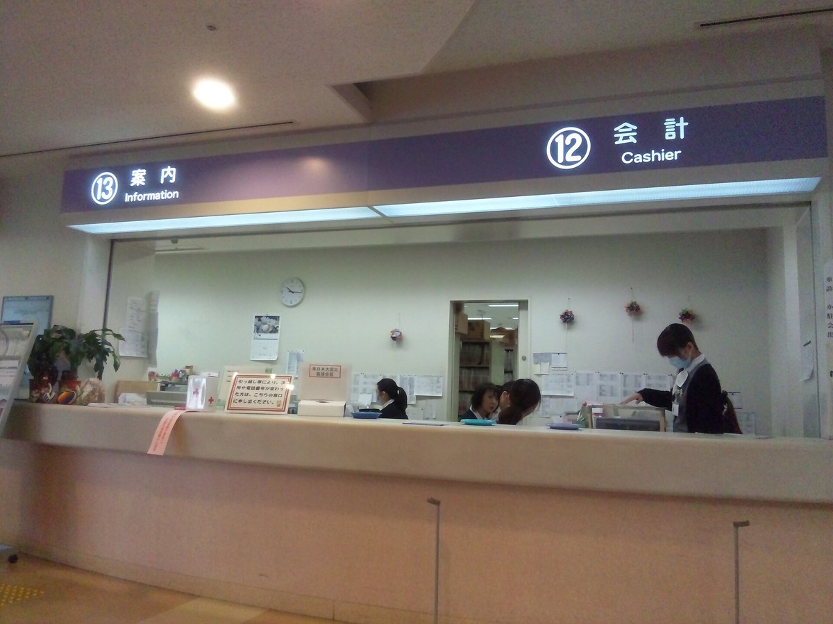 日本醫院大廳的長條櫃檯,頭頂紫色標示牌左邊寫著「⑬ 案内 Information」、右邊是「⑫ 会計 Cashier」,櫃檯後方牆面貼滿白色告示,一名戴口罩的訪客站在會計窗口前
