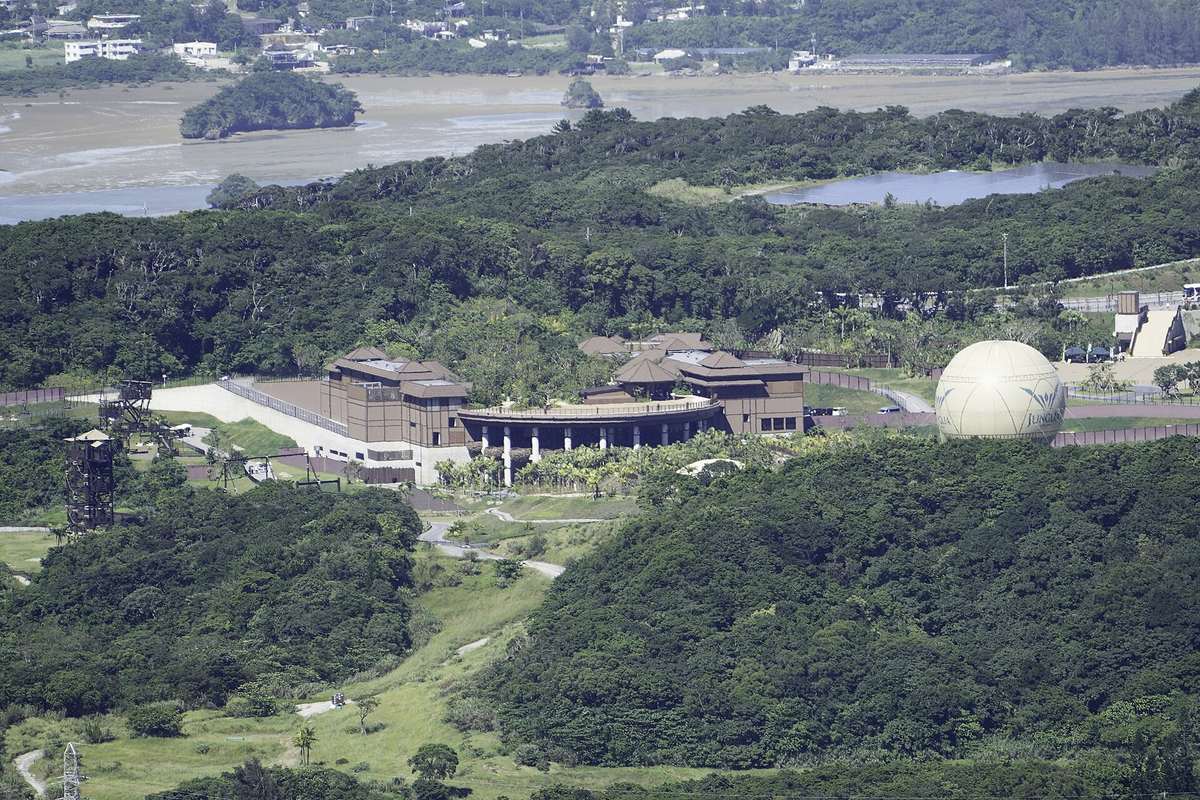 從山上遠眺沖繩本島北部的 JUNGLIA 園區:褐色斜屋頂的建築群與一排白柱廊被整片亞熱帶闊葉林包住,右側立著一顆印有 JUNGLIA 字樣的米白色巨型氣球,左邊有一座金屬瞭望塔,背景是退潮的內海與對岸聚落