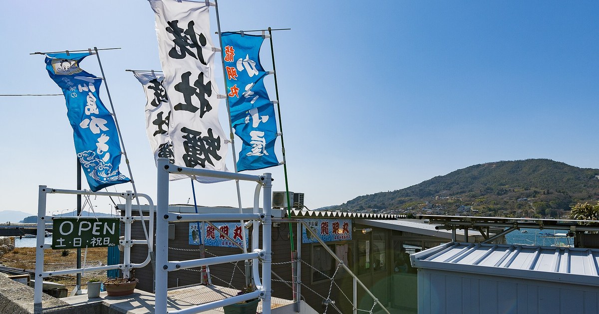 海邊一排平房前插著四面日文直幡,寫著「牡蠣」「かき小屋」等字樣;左下方一塊綠底白字的看板寫著 OPEN 與「土・日・祝日のみ」,平房門面挂著「かき小屋 龍明丸」的藍底招牌,右後方是海面、漁船與一座住宅散布的山丘