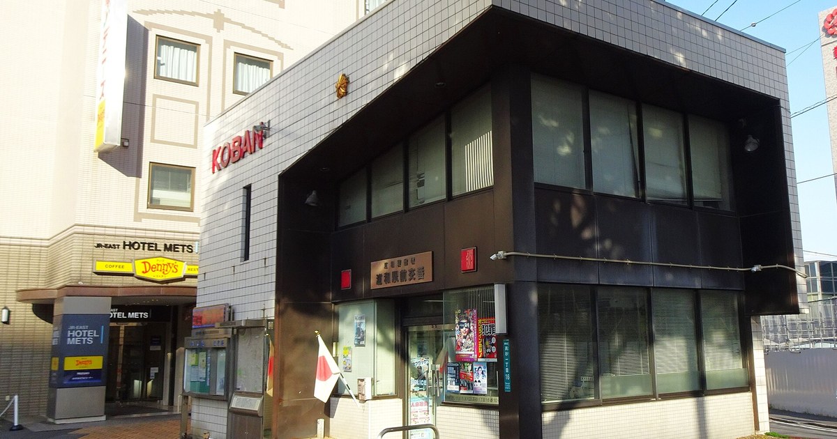 日本浦和站前的交番,白色磁磚外牆上有紅色立體字母 KOBAN,深色雨庇下掛著銅色門牌與金色警徽,門口立著日本國旗,玻璃門旁貼滿通緝與宣導海報,左側是 JR-EAST HOTEL METS 與 Denny's 的招牌