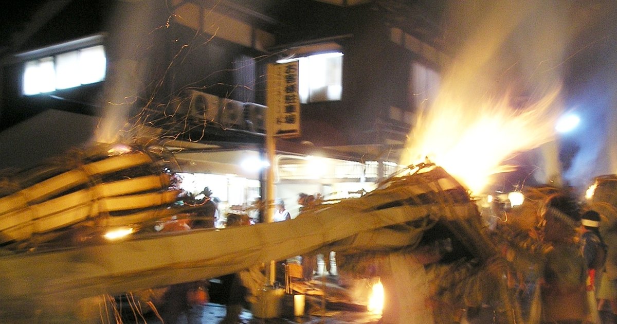 夜裡的鞍馬聚落,燒著的大松明被扛著橫過街道,火焰和火星往上竄,後方是民家與路燈