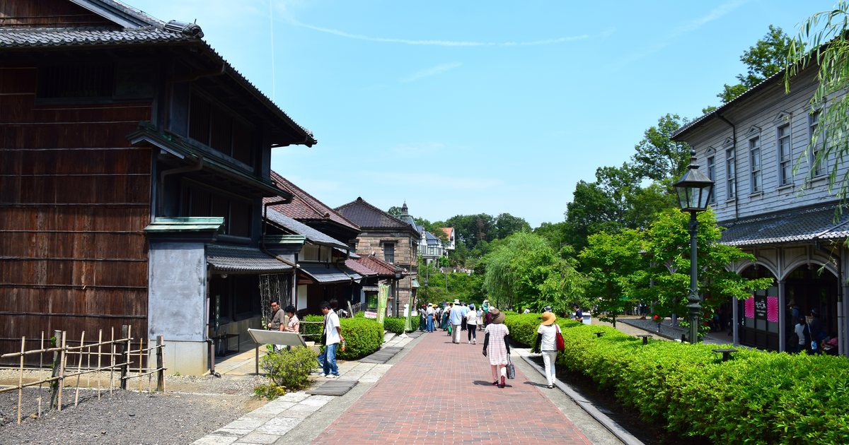 博物館明治村的紅磚步道，左側是深色木造的日式建築、右側是白色木造的西洋式二層樓，中間有煤氣路燈與往深處走去的遊客