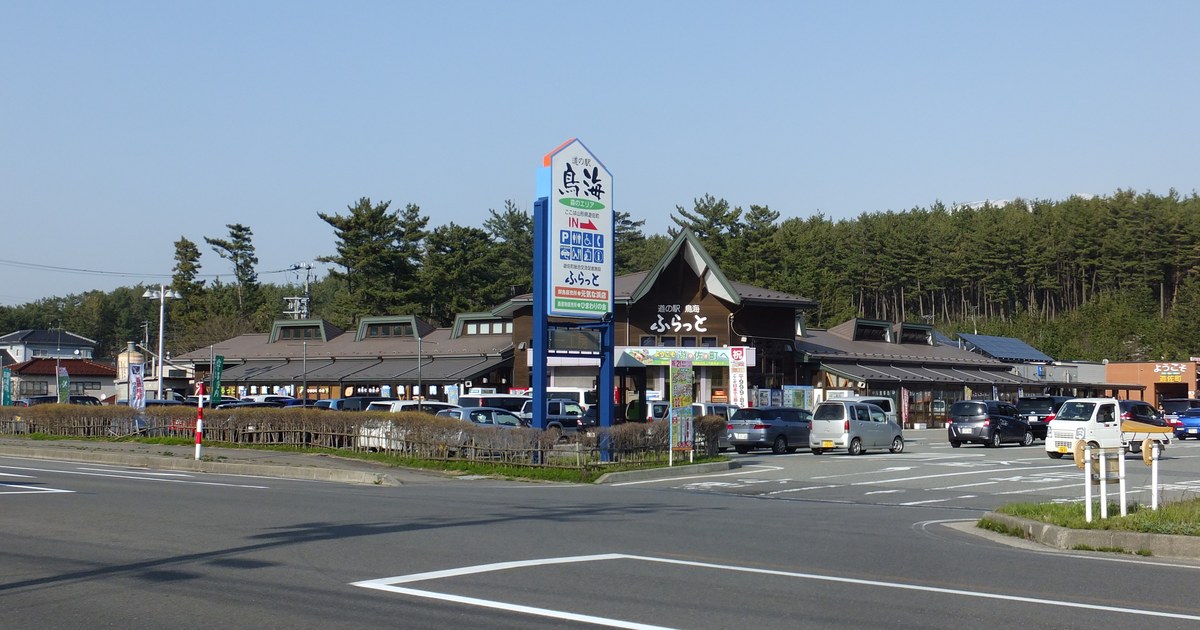 道の駅鳥海「ふらっと」的站區外觀,褐色斜屋頂的低矮站舍前停滿轎車與輕型車,藍色高柱招牌標示入口與停車、廁所、餐飲圖示,前景是國道路面。
