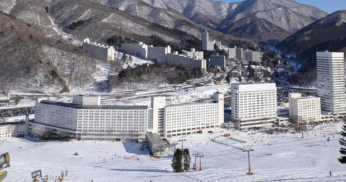 苗場滑雪場山腳：一整排白色的苗場王子飯店大樓沿著雪場底部排開，右側是一棟高塔樓，前景是壓雪雪道、吊椅纜車支柱和造雪機，後方是覆雪的山谷與成排公寓大樓