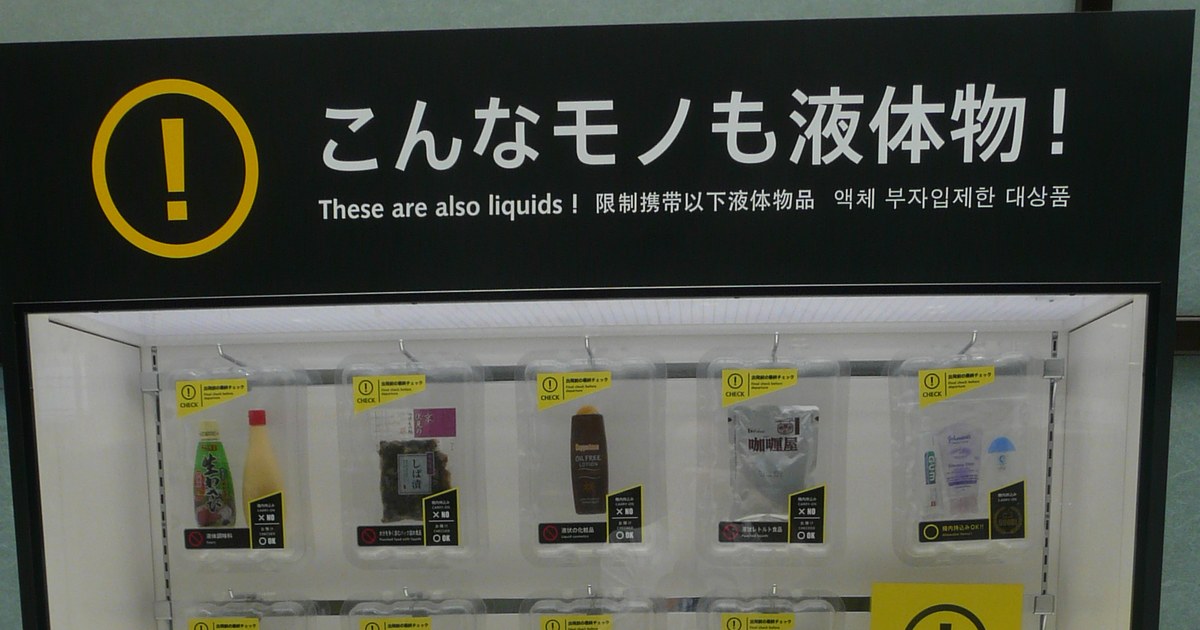 成田機場的液體物展示櫃:黑色頂板上印著黃色驚嘆號與「こんなモノも液体物! These are also liquids!」,四種語言並列;玻璃櫃內第一排掛著五個透明托盤,分別裝著液體調味料、しば漬醬菜、化妝水、咖哩調理包,以及一袋裝了牙膏與乳液的透明夾鏈袋。