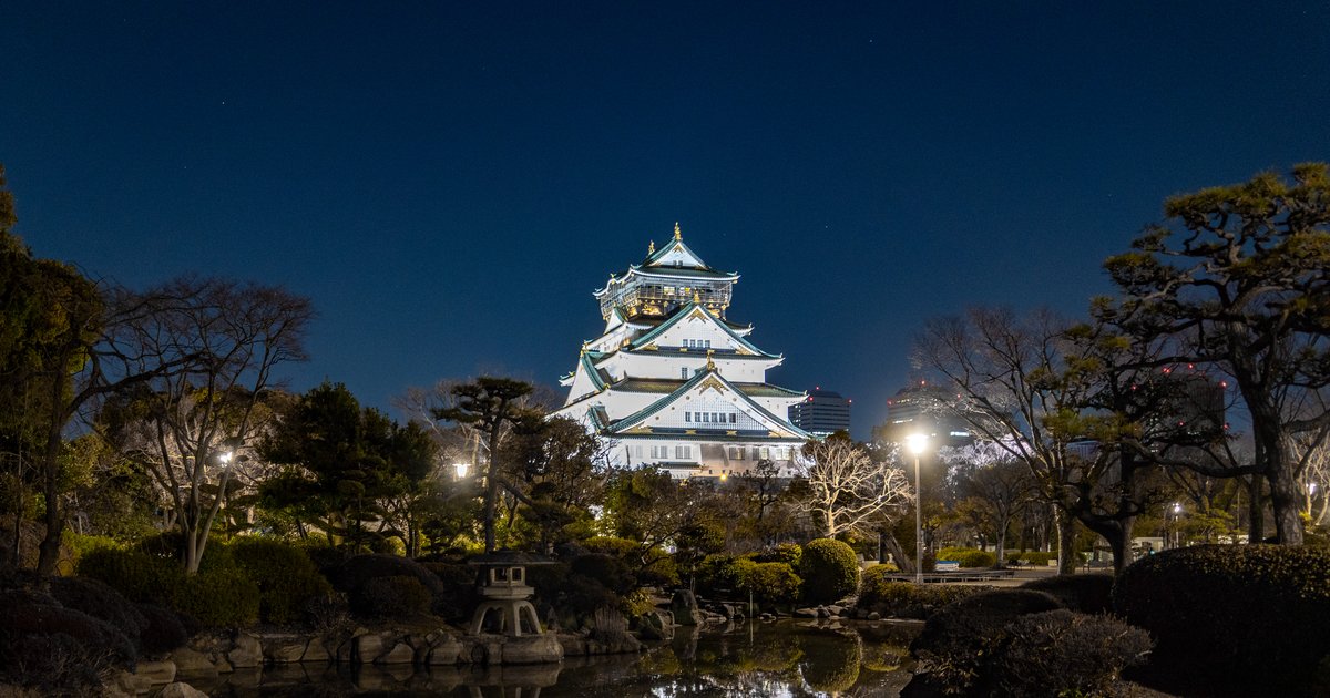2 月晚上的大阪城天守閣，白色外牆與綠色屋頂整棟打亮，前景是本丸日本庭園的水池、石燈籠和落葉的樹，池面映出天守閣的倒影