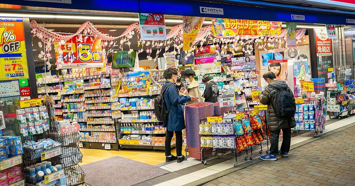 日本藥妝店的開放式店面:騎樓底下三面貨架從地板排到頭頂,滿是藥品、日用品與化妝品的紙盒,天花板垂掛粉色櫻花裝飾與黃色特價布條,右側門柱掛著「免税店 Tax-Free Shop」看板,幾名戴口罩的顧客站在架前挑東西,門口疊著一整落購物籃。