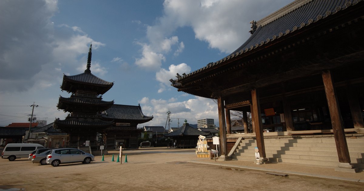 西大寺観音院的境內廣場:右側是本堂,深灰瓦頂與翹起的簷角、正面一排粗大的木柱與石階;左側遠處是三重塔,中間是一大片空曠的沙土地,幾輛車停在左邊,天空有大片積雲