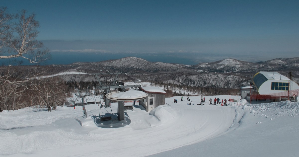 札幌国際スキー場 山頂晴天雪景,吊椅纜車頂站的圓形滑輪罩與小屋、雪坡上一群滑雪客,遠處是層層覆雪山稜與地平線上的一片海面