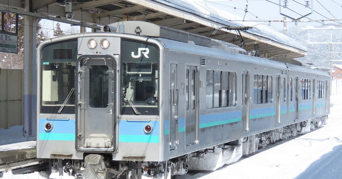 冬天積雪的白馬站月台旁,停著一列車頭有 JR 標誌、銀色車身配藍綠色腰帶的 E127 系普通電車,後方是覆雪的山坡
