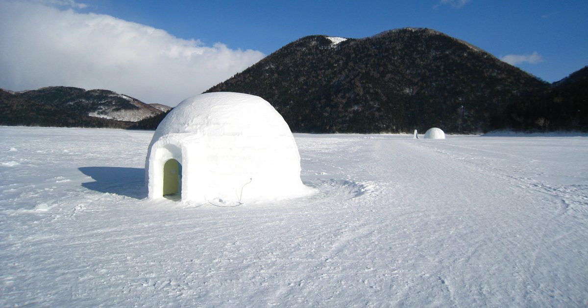 結冰的然別湖湖面上一棟雪塊砌成的圓頂冰屋,入口朝左,遠處湖面還有另一棟小冰屋,背後是覆著樹林的天望山與藍天