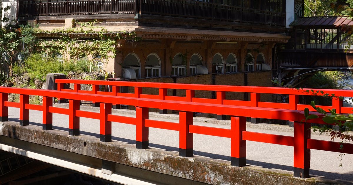 四万川上的慶雲橋：紅色木欄杆橫過整個畫面，橋後面是積善館本館底層那一排半圓拱窗與爬滿藤蔓的石造基座，再上面是木造二樓的黑色迴廊