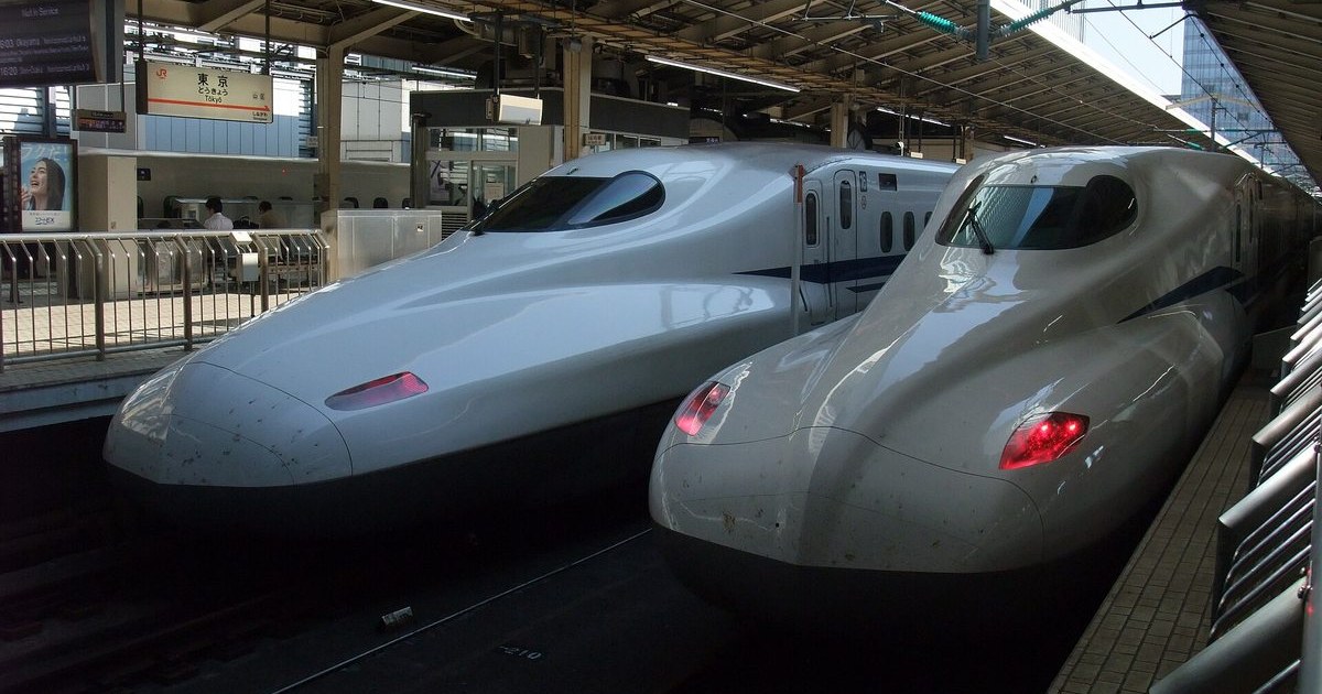 東京車站新幹線月台上兩列並排停靠的白色新幹線，鴨嘴形車頭、車身有藍色腰帶，右側那列的尾燈亮著紅色；左上角是「東京 Tokyo」的 JR 站名牌與發車資訊看板