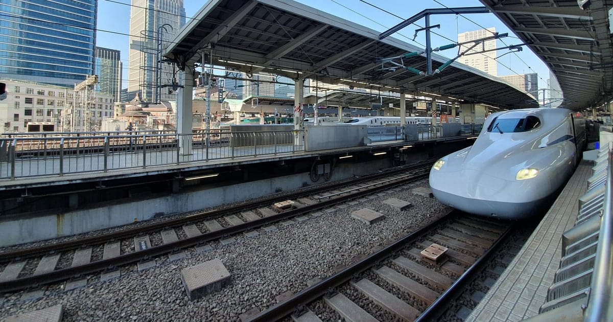 東京車站的東海道新幹線月台,一列白色 N700S 系列車的車頭正沿著彎道駛入,左側是月台雨棚與丸之內側的高樓群,天空晴朗無雲