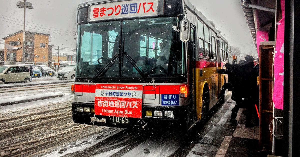 大雪中的十日町雪まつり巡迴巴士,車頭 LED 顯示「雪まつり巡回バス」,前保險桿掛著寫有「十日町雪まつり 市街地巡回バス」的紅色看板,車門邊有乘客正在上車