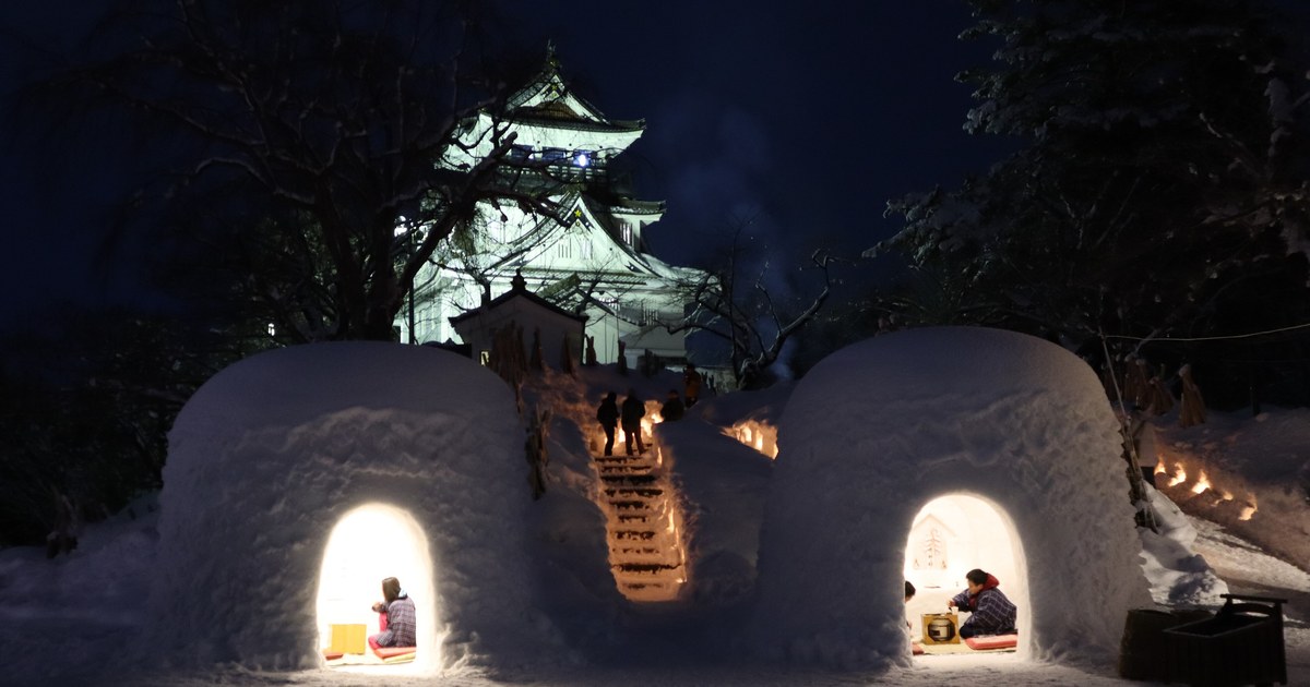 夜間的橫手公園：兩座雪屋亮著暖黃燈光、裡面各坐著孩子，中間是插滿蠟燭的雪階，後方點燈的橫手城天守