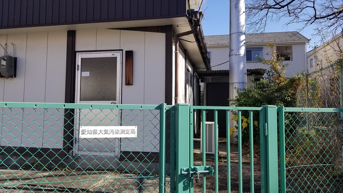 住宅區裡一棟白色小平房，門旁掛著寫有「愛知県大気汚染測定局」的牌子，屋外立著一根高過屋頂的不鏽鋼採樣管，整棟被綠色鐵絲網圍籬與上鎖的門圍住