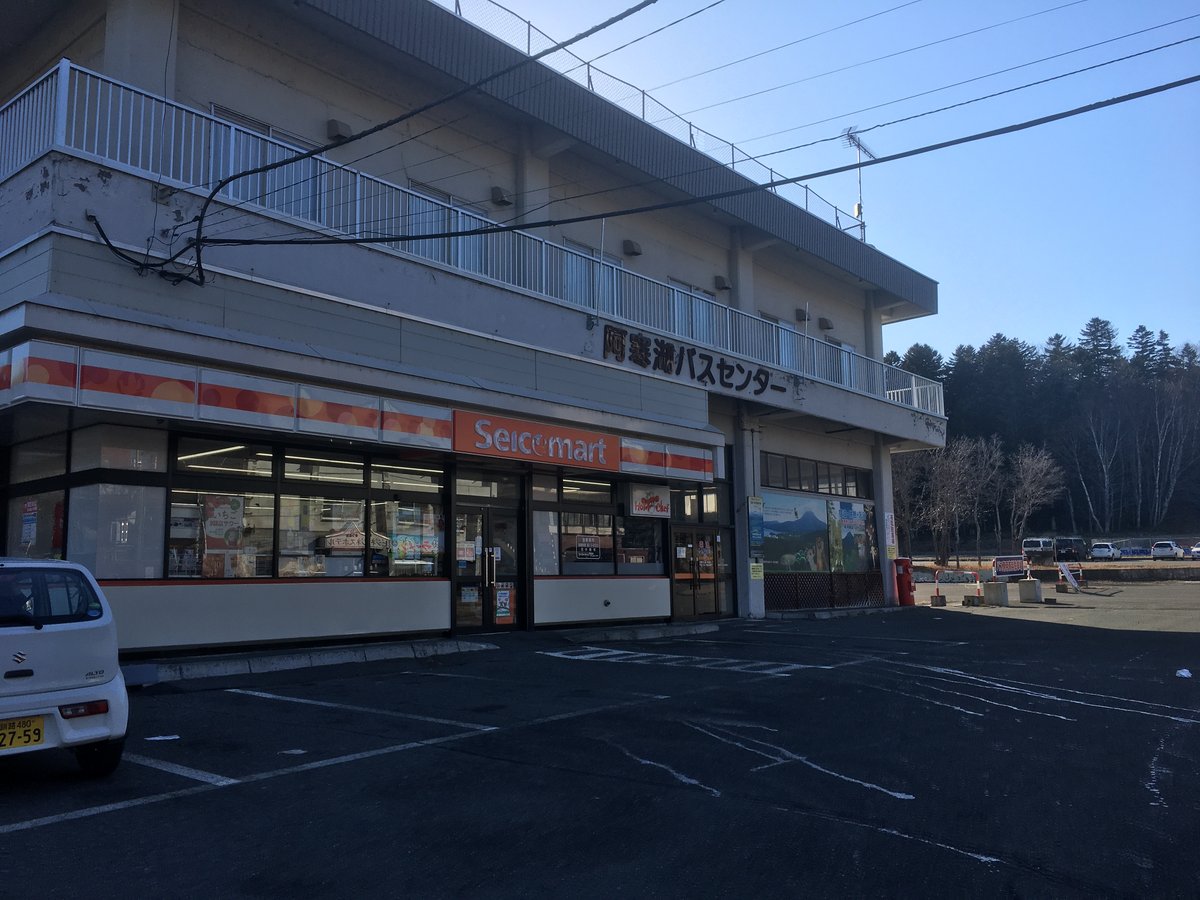 阿寒湖巴士中心的外觀:兩層樓的米白色建築,二樓外牆掛著「阿寒湖バスセンター」的黑色字,一樓是橘色招牌的 Seicomart 便利商店,門前是一片畫好停車格的柏油廣場