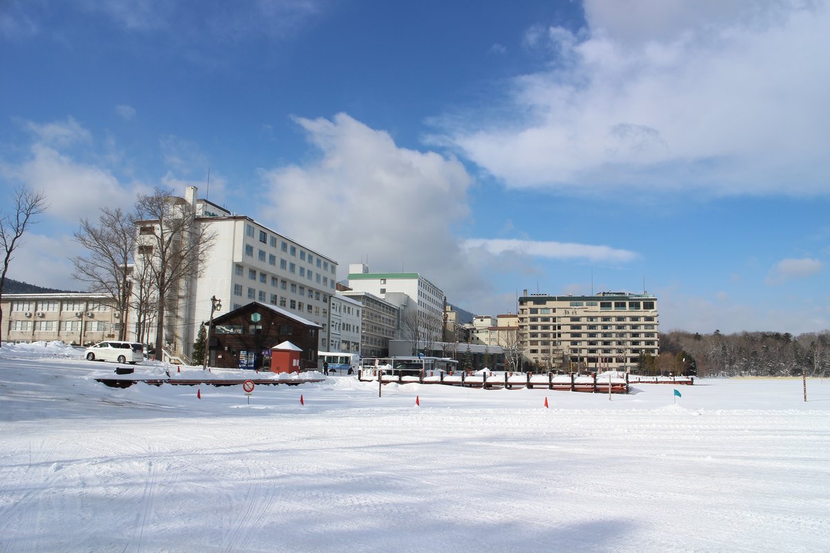 2 月的阿寒湖溫泉:前景是整片結冰積雪的湖面,雪地上插著一排橘色標示旗和一面禁止進入的圓形標誌,中景的木造棧橋被雪埋住一半,後方一整排是阿寒湖溫泉街的多層旅館建築