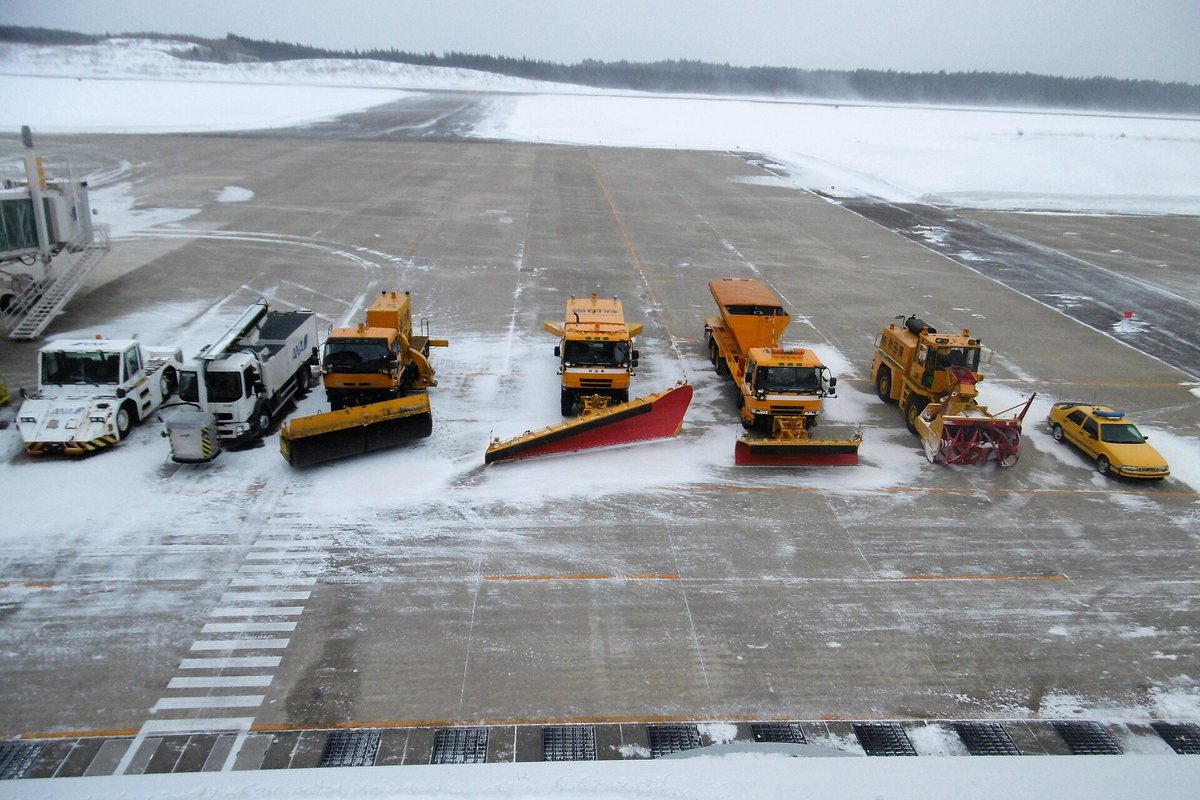秋田・大館能代空港停機坪上一字排開的除雪車隊，包含推雪板卡車、撒布車與鼓風式除雪機，遠方跑道飄著雪