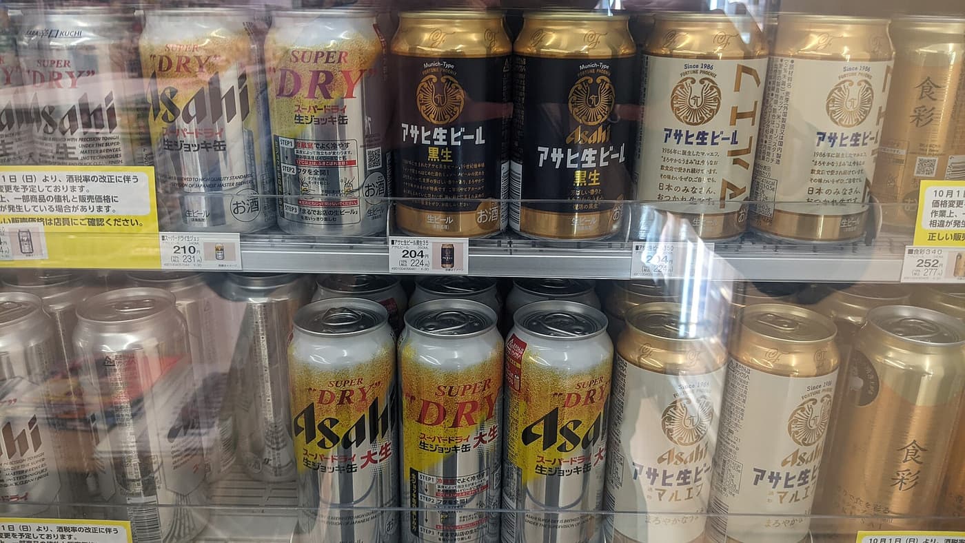 日本超商的啤酒冷藏櫃內部特寫:兩層架上並排著 Asahi スーパードライ生ジョッキ缶、アサヒ生ビール黒生與マルエフ 等罐裝啤酒,架緣夾著白色價標寫著 210 円與稅込 231 円,貨架左右兩端各貼著一張黃色告示,寫著 10 月 1 日起因酒稅率改正而預定變更價格