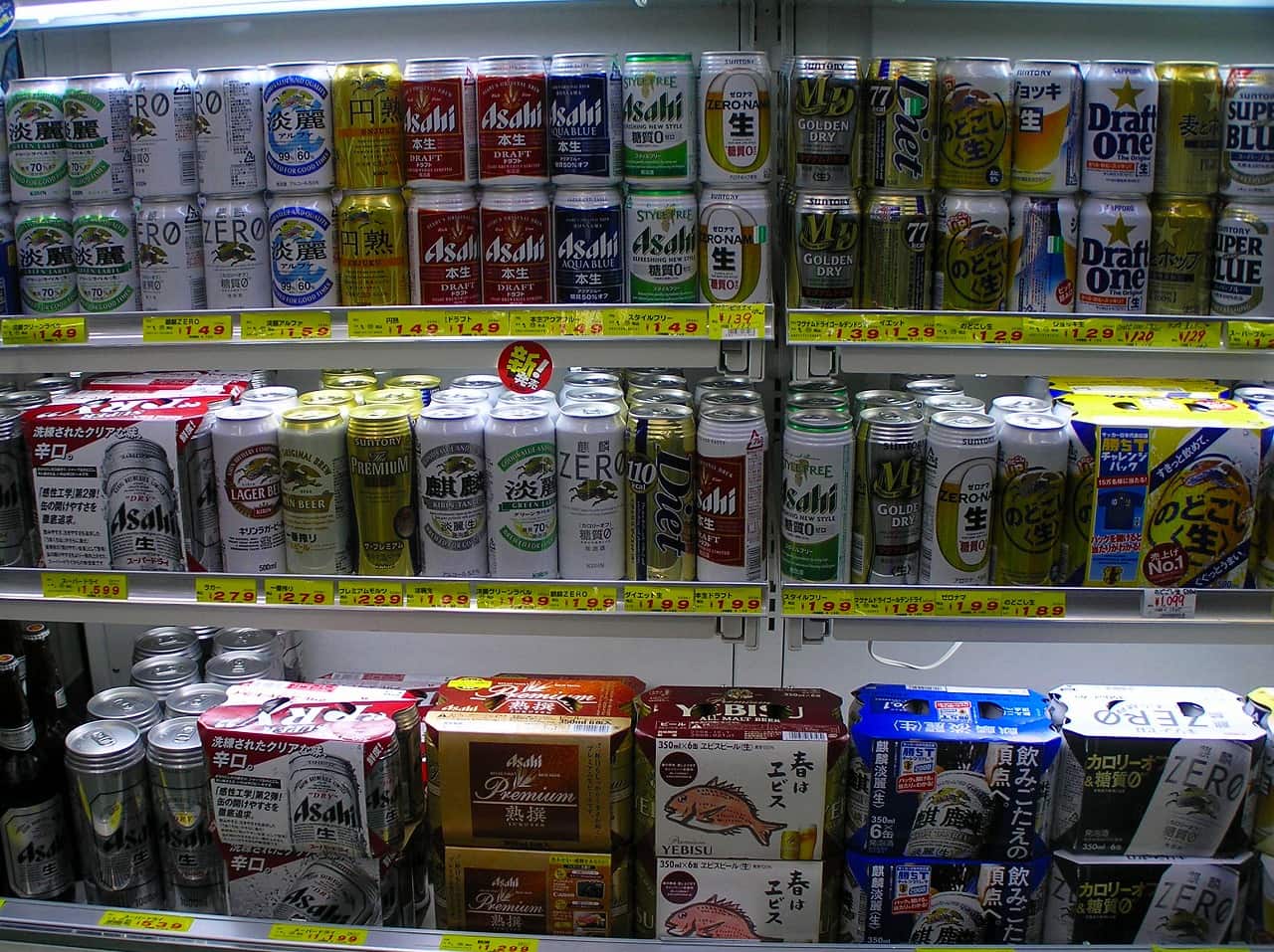 日本量販店的啤酒貨架三層特寫:上層與中層整排立著 350 毫升鋁罐,包含麒麟淡麗、アサヒ本生、スタイルフリー、サッポロ Draft One、のどごし〈生〉等,每一格下緣夾著黃色手寫價標,價格從 120 到 299 円不等,最下層是六罐裝紙盒