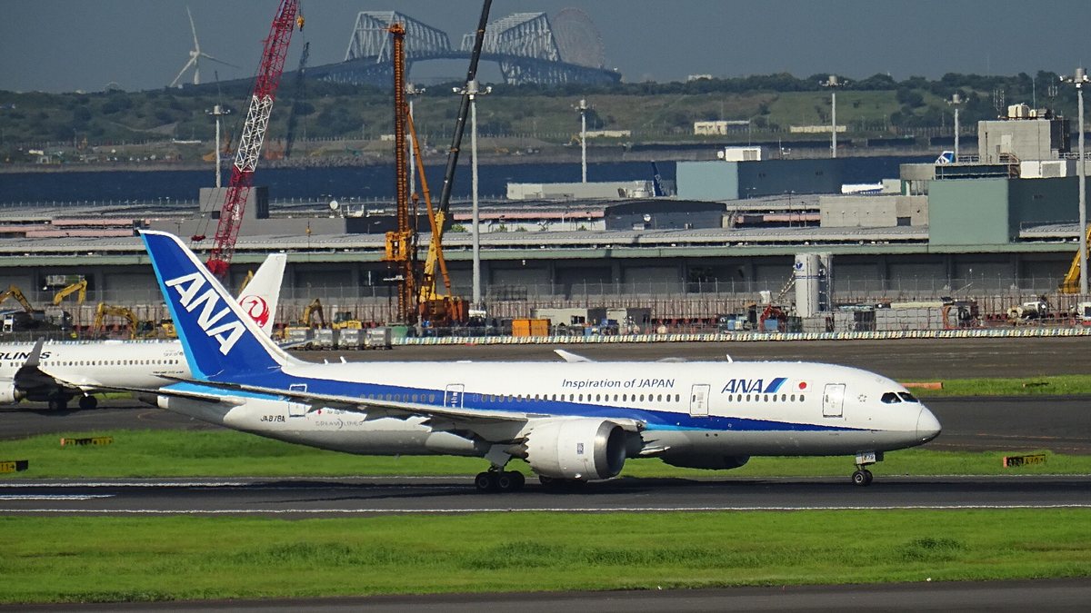 ANA 波音 787-8 在羽田機場跑道上加速,藍色垂直尾翼、機身寫著 Inspiration of JAPAN 與註冊編號 JA878A,左後方露出一架日本航空的紅色鶴丸尾翼,遠處是東京門橋