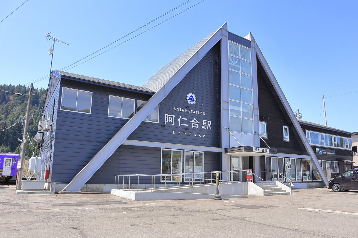 阿仁合站站體：深藍色的三角屋頂建築，白色大字寫著「ANIAI-STATION 阿仁合駅 しあわせのえき」，右邊是玻璃樓梯間，入口有無障礙斜坡與階梯，左邊停著一列紫白色的秋田內陸線列車，站前是空曠的柏油地