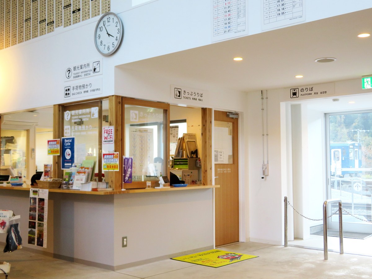 阿仁合站站內：左邊是掛著「観光案内所・手荷物預かり」牌子的木質櫃台，中間木框窗口上方寫著「きっぷうりば TICKETS」，右邊門口標示「のりば PLATFORM」，玻璃門外停著一列內陸線列車，牆上有圓形時鐘與幾張時刻表