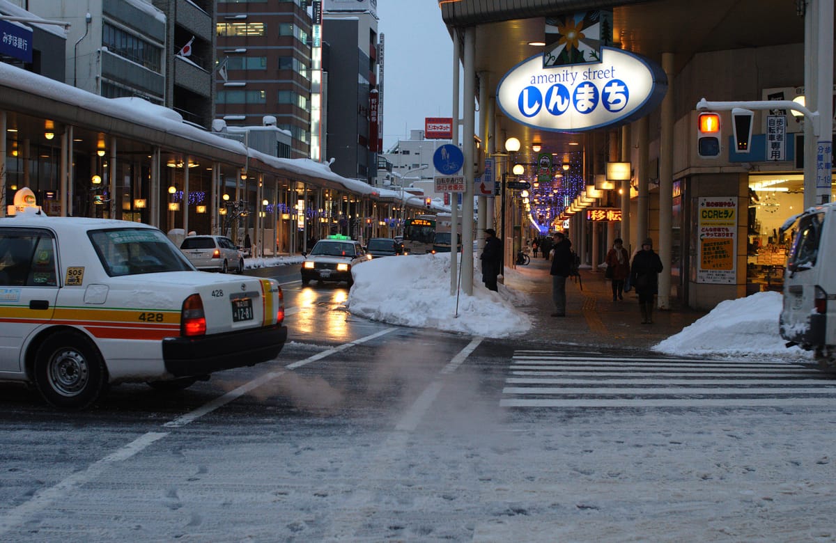 1 月中旬青森市新町通的街景,車道被壓實的雪覆蓋,人行道與車道之間堆著除雪堆出的雪牆,計程車與行人在雪中通行