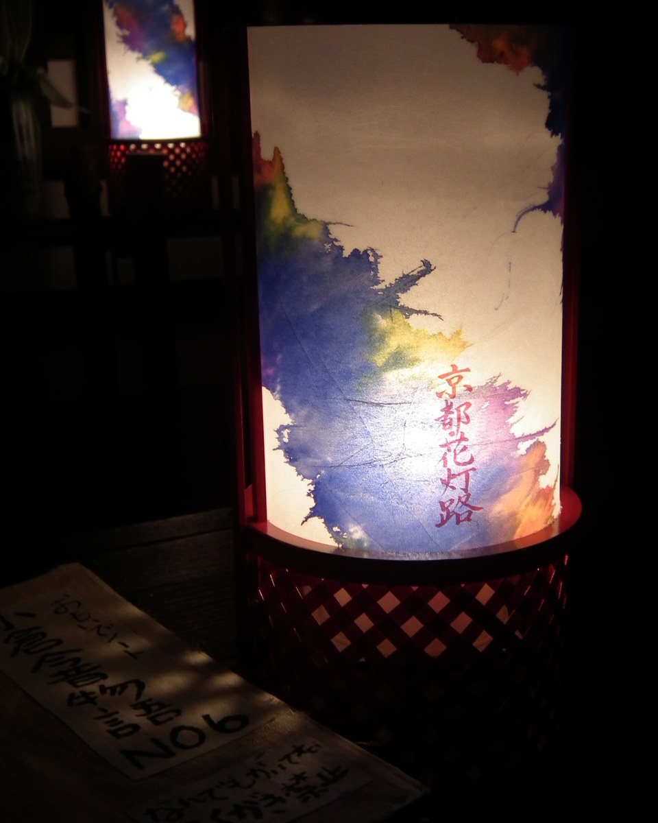 夜裡點亮的花燈路行燈特寫:圓筒和紙燈罩上有藍紫色暈染花紋,印著紅色「京都花灯路」字樣,後方還有一盞同款行燈