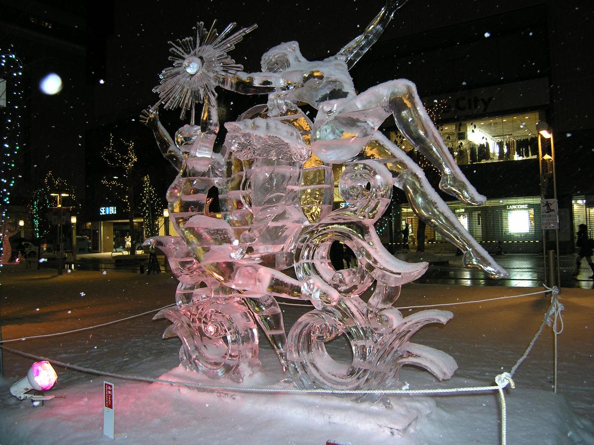 下雪的夜裡,平和通買物公園步行街上一座打粉紅燈的冰雕,雕著舉著太陽的人物與浪花,四周拉繩圍起,背後是西武百貨與街邊商店的燈光